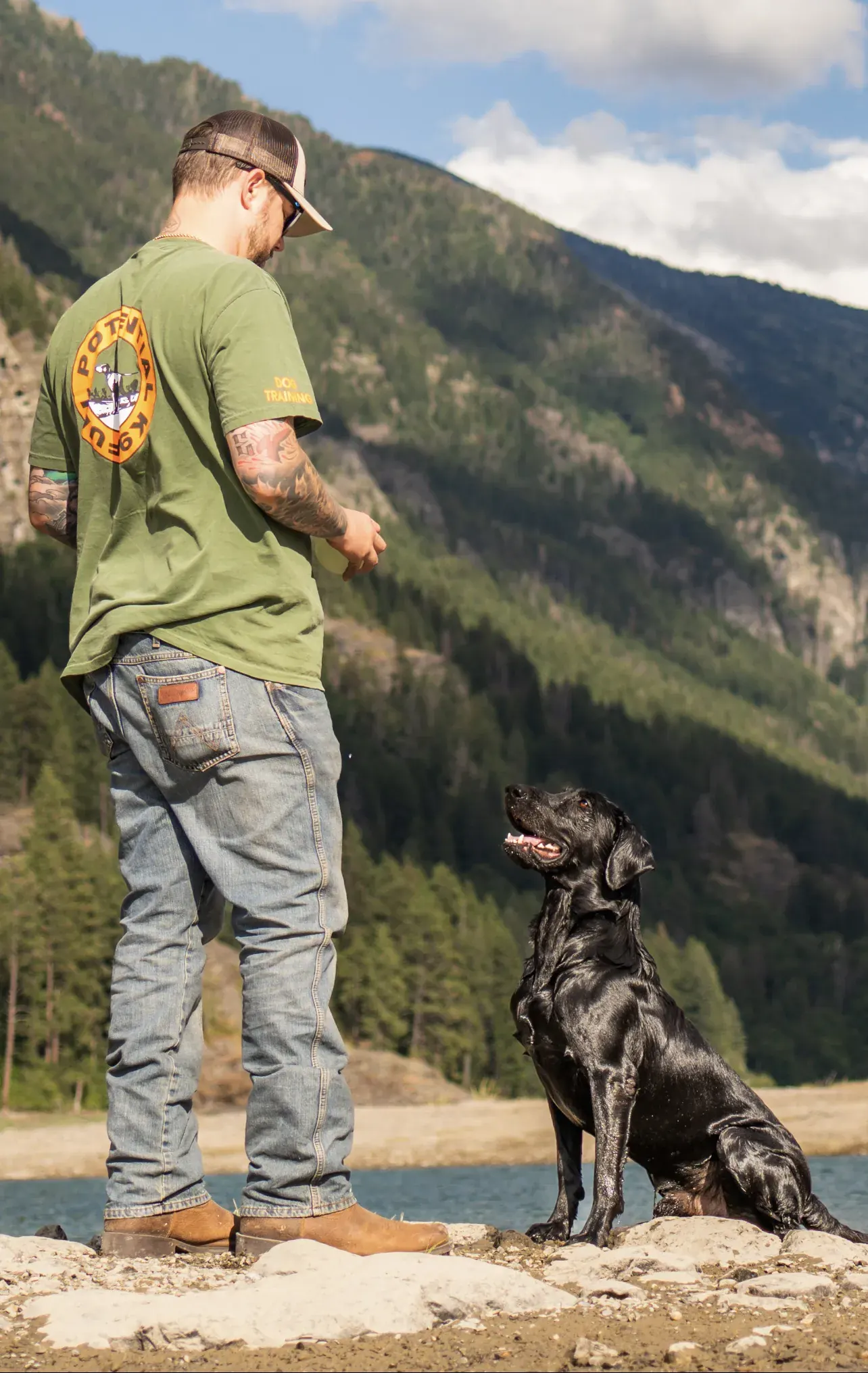 Dillon Mitchell standing on rocks beside a lake as a black Labrador sits and looks up at him, waiting for a cue