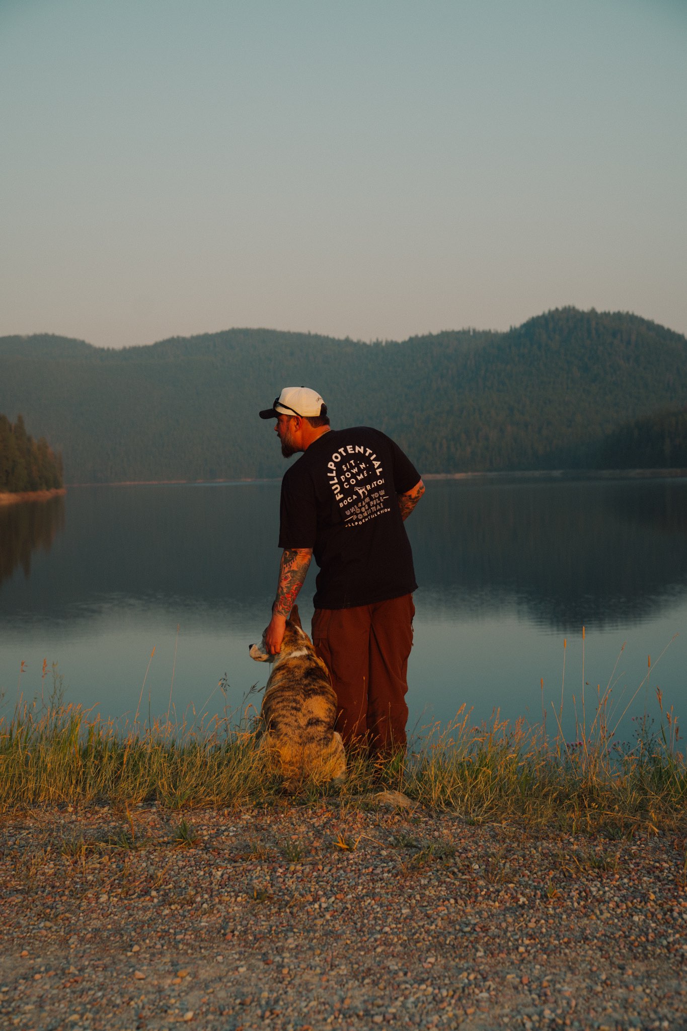 Founder Dillon Mitchell in a Full Potential K9 shirt overlooking a northwestern Montana mountain lake at sunset with a trained dog