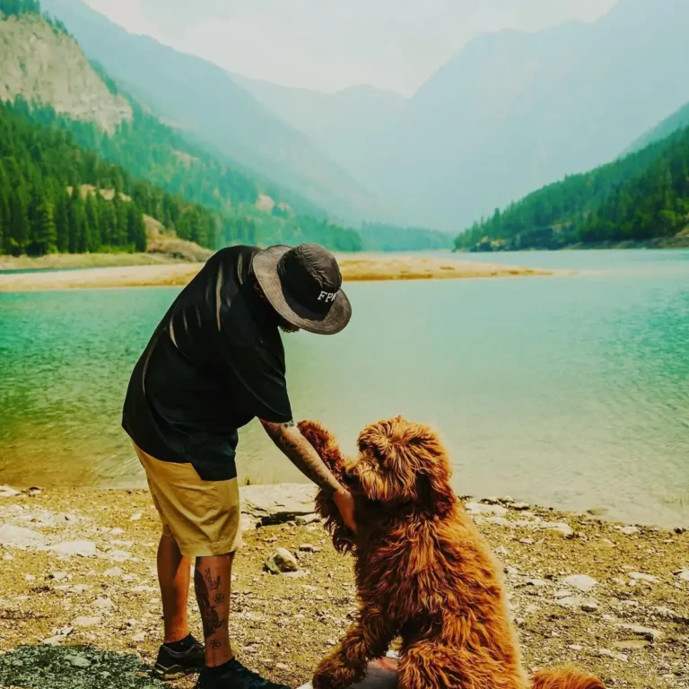 A Full Potential K9 trainer in a bucket hat shaking paws with a curly red doodle on a rocky mountain lakeshore