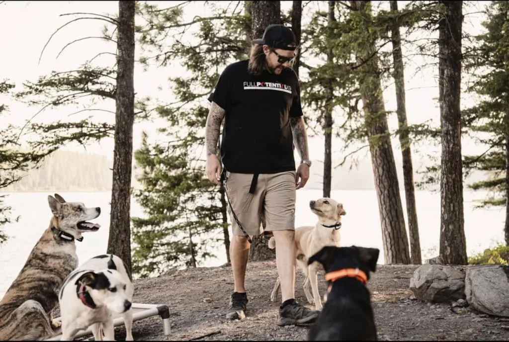 A Full Potential K9 trainer walking calmly with several dogs along a forested lakeshore